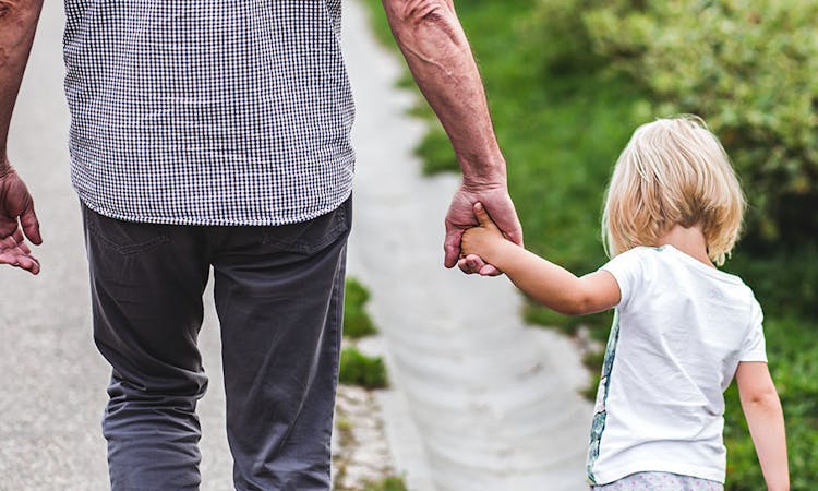 Image of an older man and younger child holding hands, taken from the back. The man is wearing a checkered shirt and grey trousers, and the young girl is wearing a white tshirt. They are walking in the countryside, you can see greenery in front of them.