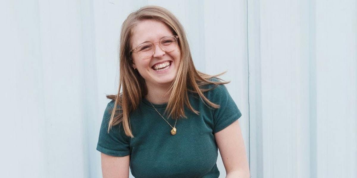 Image of our procurement manager, Ellie, stood in front of a white wooden slated wall. She's wearing a green t-shirt and is mid laugh, holding her hands together in front of her waist.