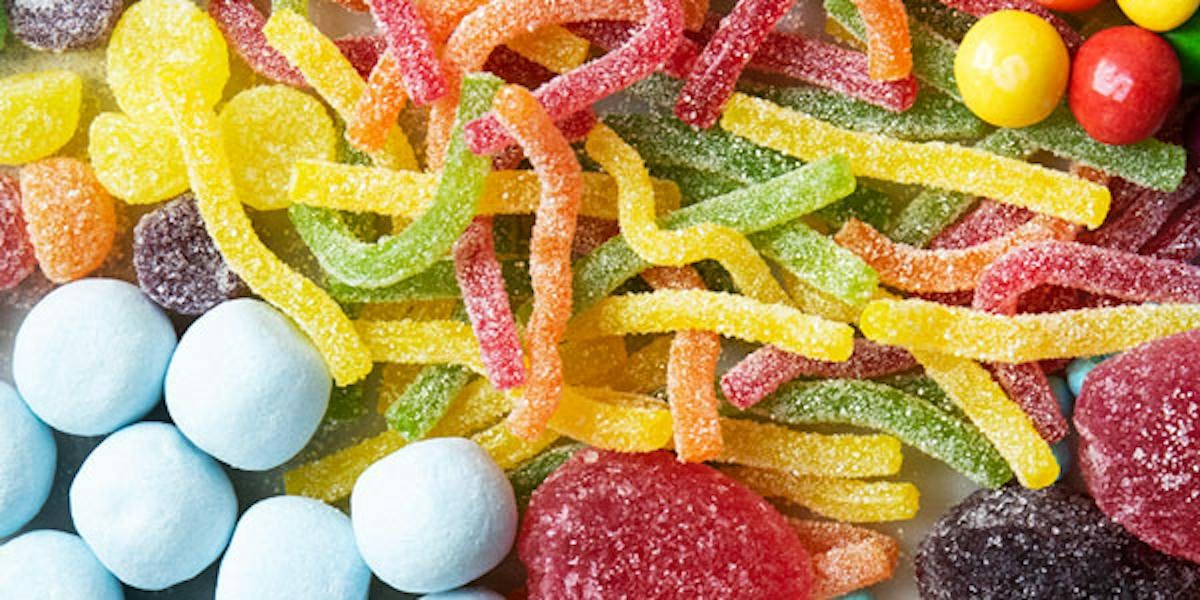 Close up image of sweets layed on a surface. There are fizzy worms in the middle, skittles in the top right, bonbons in the bottom left, jelly fruits in the bottom right, and fizzy jelly fangs in the top left.