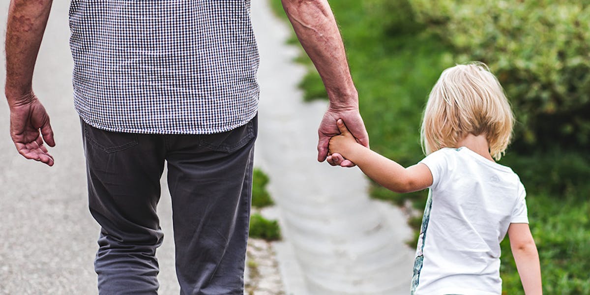 Image of an older man and younger child holding hands, taken from the back. The man is wearing a checkered shirt and grey trousers, and the young girl is wearing a white tshirt. They are walking in the countryside, you can see greenery in front of them.