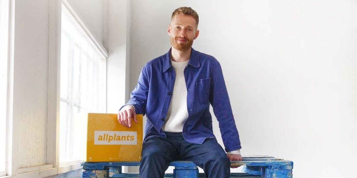 Image of our director of customer experience, Ferdie, sat on top of a stack of blue palettes wearing slack trousers, a white t-shirt, and an unbuttoned blue shirt. He's smiling with his left arm resting on an allplants delivery box.